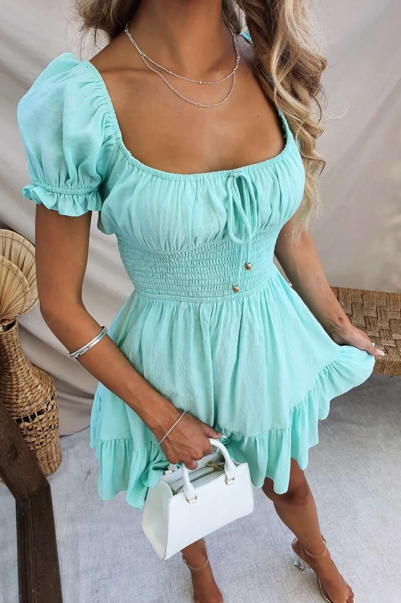 Model wearing Love In Motion Dress in mint green, featuring a tie front and elastic waistband, paired with a white handbag.