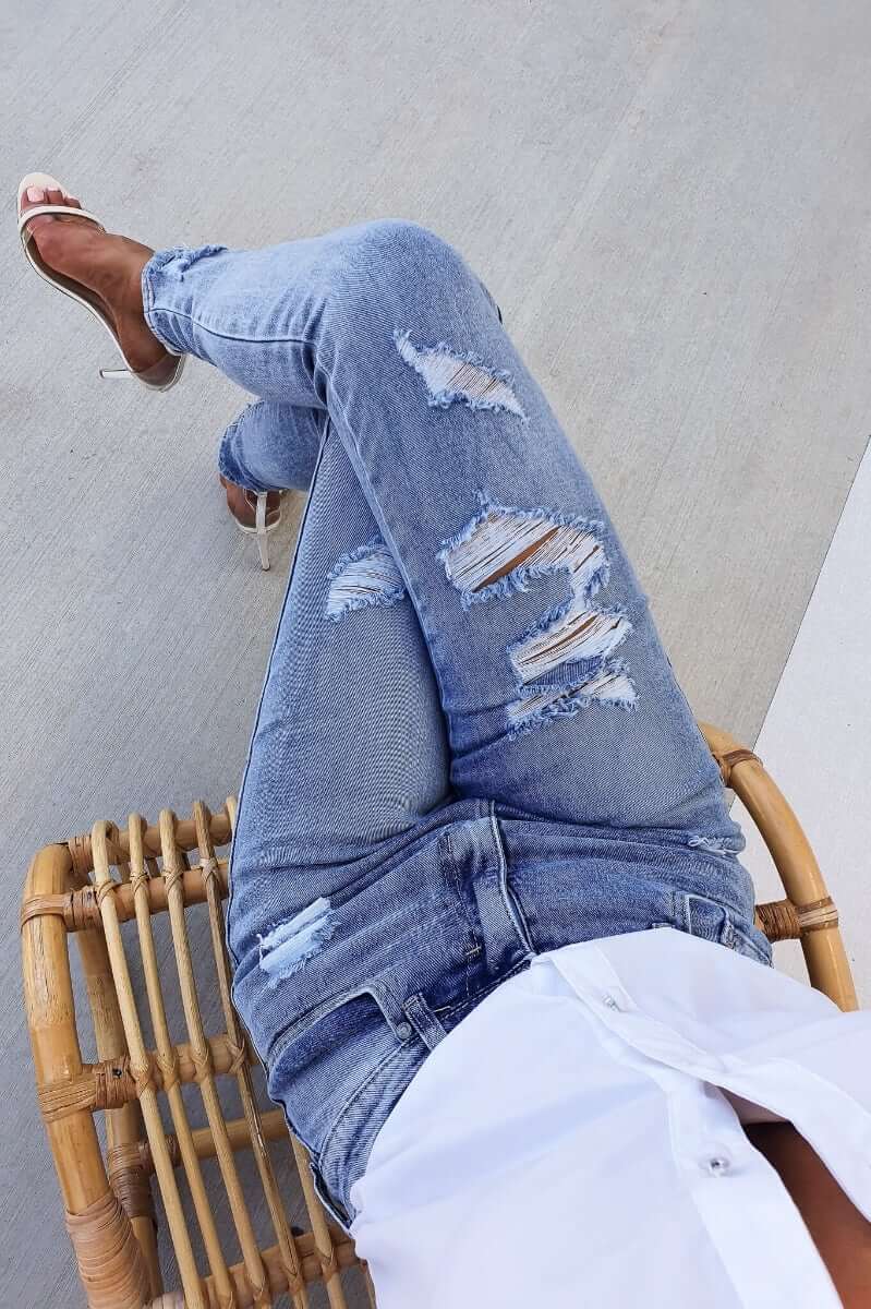 Mecka Jeans in distressed blue denim with mid-high waist fit and pockets, styled with a white shirt and strappy heels.