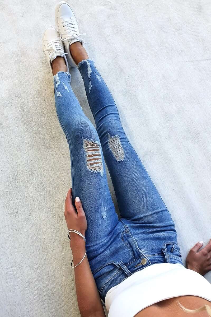 Qilli distressed denim jeans with rip details, raw hem, and stretch material, styled with a white crop top and sneakers.