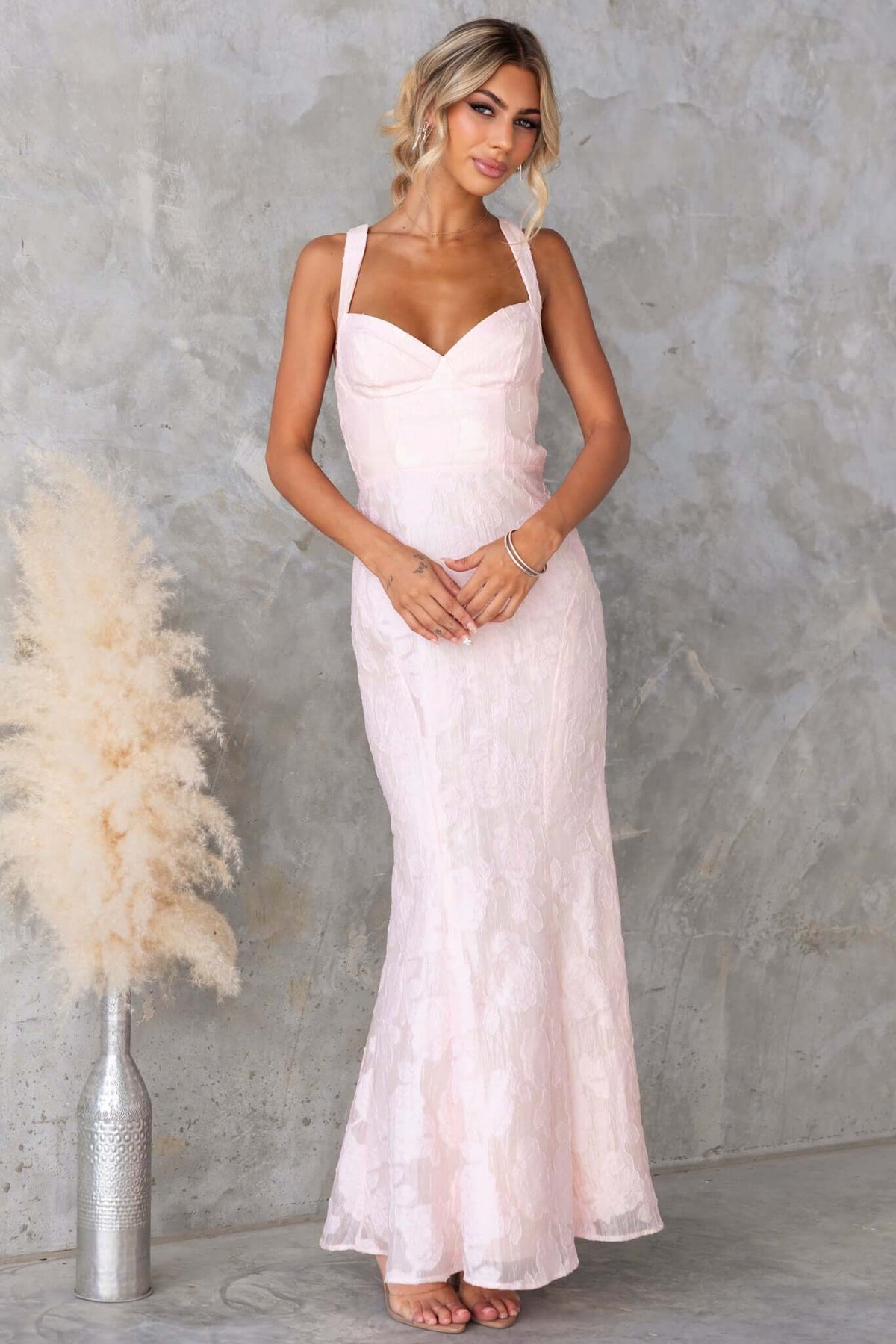 Remma Maxi Dress in soft pink with lace-up back and slight fishtail hem, showcased by a model on a textured backdrop.