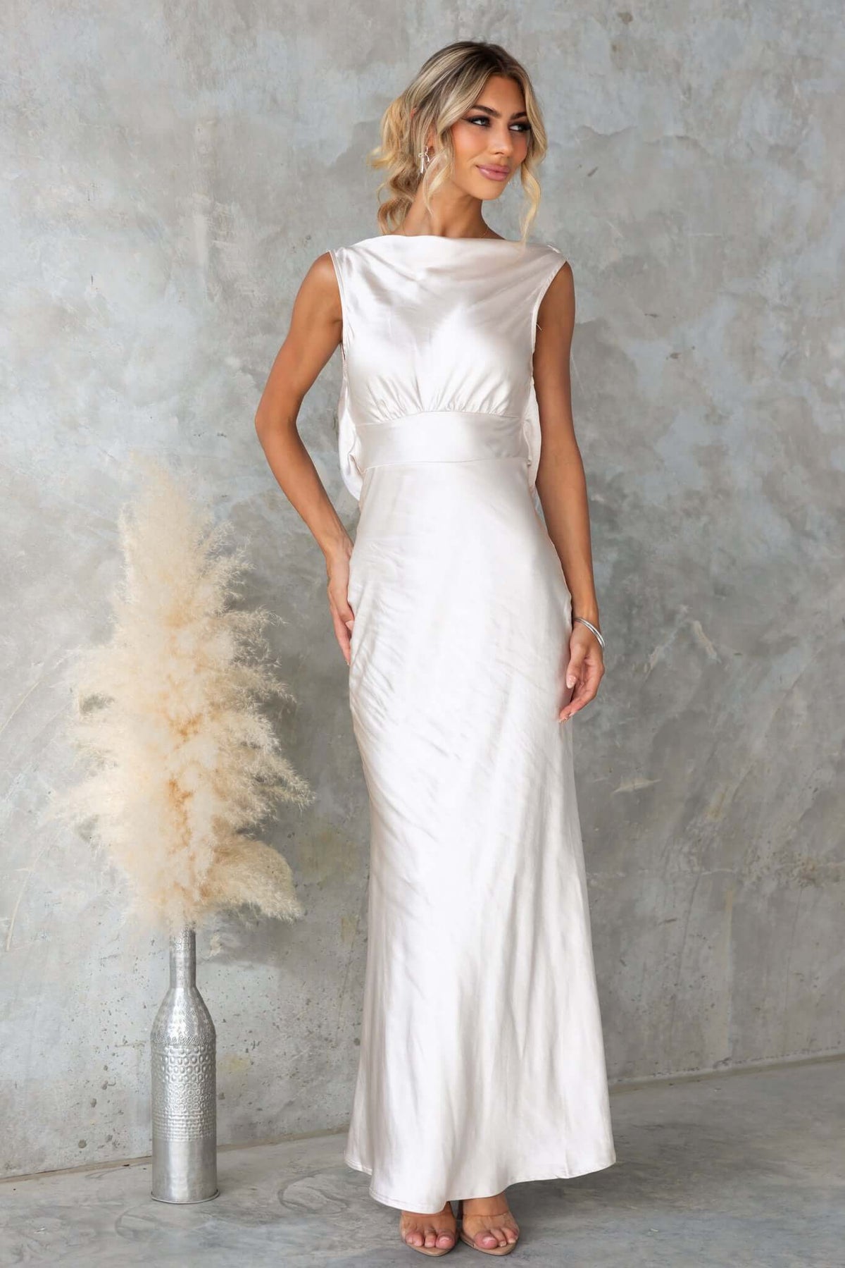 Silvia Maxi Dress in satin with boat neckline, elegant silhouette, worn by model against a textured background.