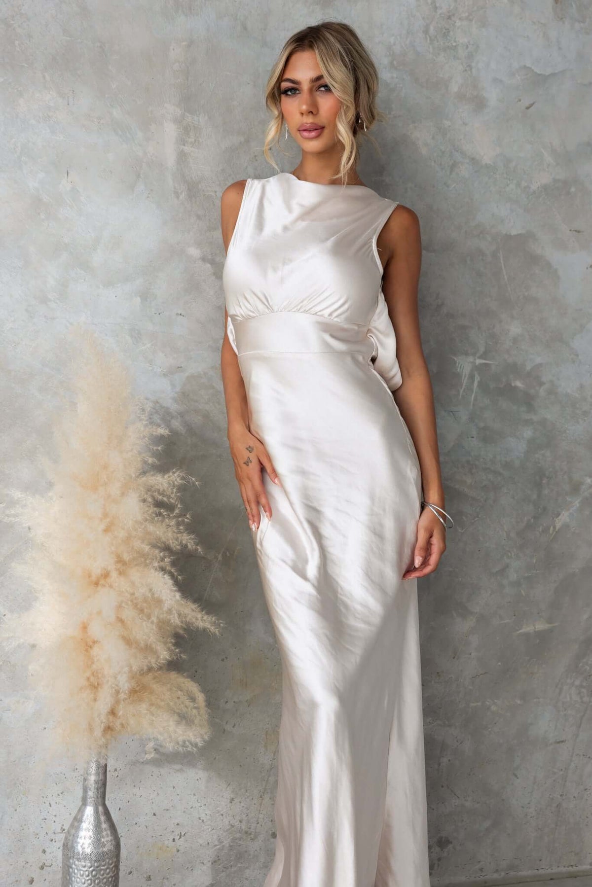 Silvia Maxi Dress featuring a boat neckline, satin fabric, and elegant draping on a model against a textured backdrop.