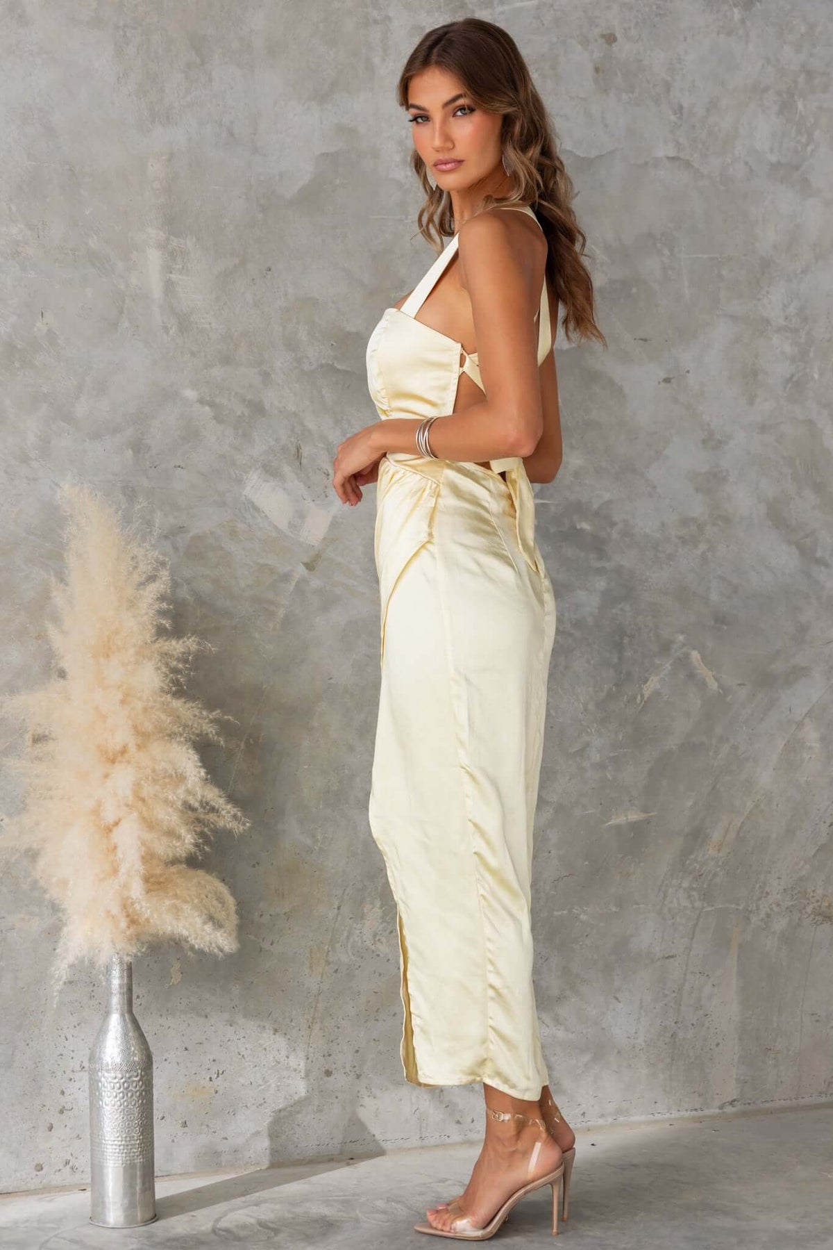 Side view of a woman wearing a yellow Merritt Maxi Dress with adjustable tie straps, styled with heels and a floral accent.