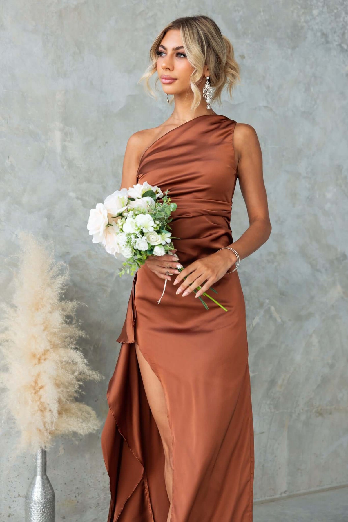 Model wearing Dimmi Maxi Dress, a satin one-shoulder gown with a high slit, holding a bouquet of flowers.
