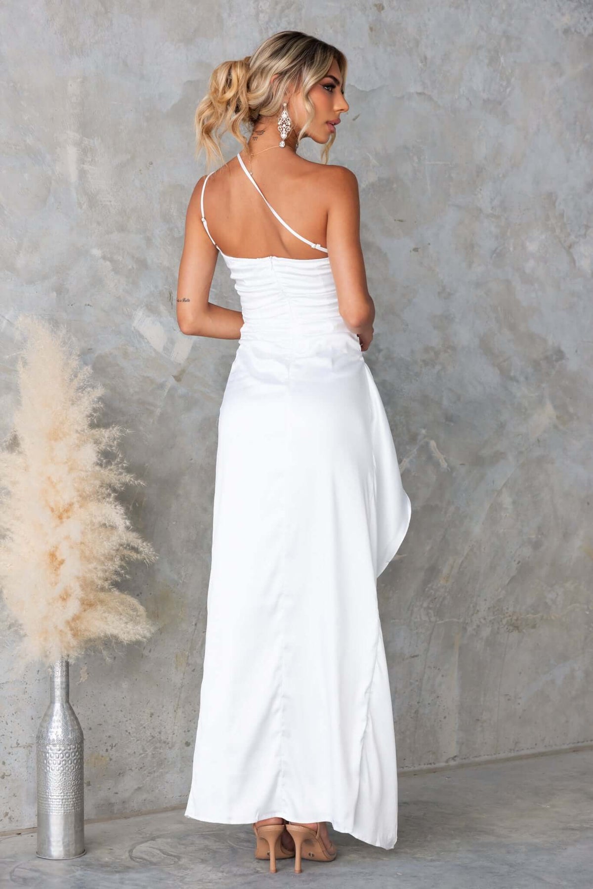 Back view of woman in white satin Dimmi Maxi Dress featuring one-shoulder neckline and ruffled hem.