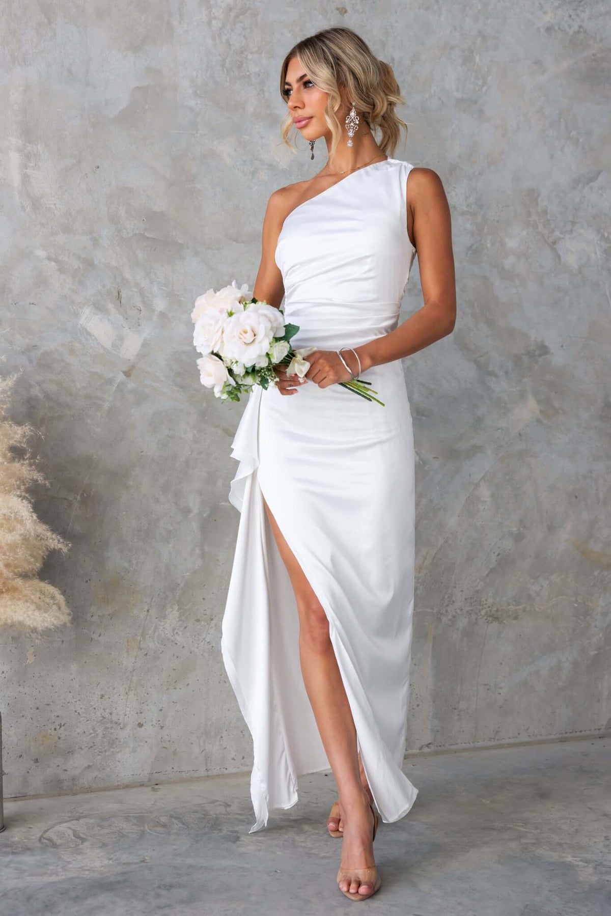 Model in a fitted white satin one-shoulder maxi dress with a ruffle trim and side slit, holding flowers.