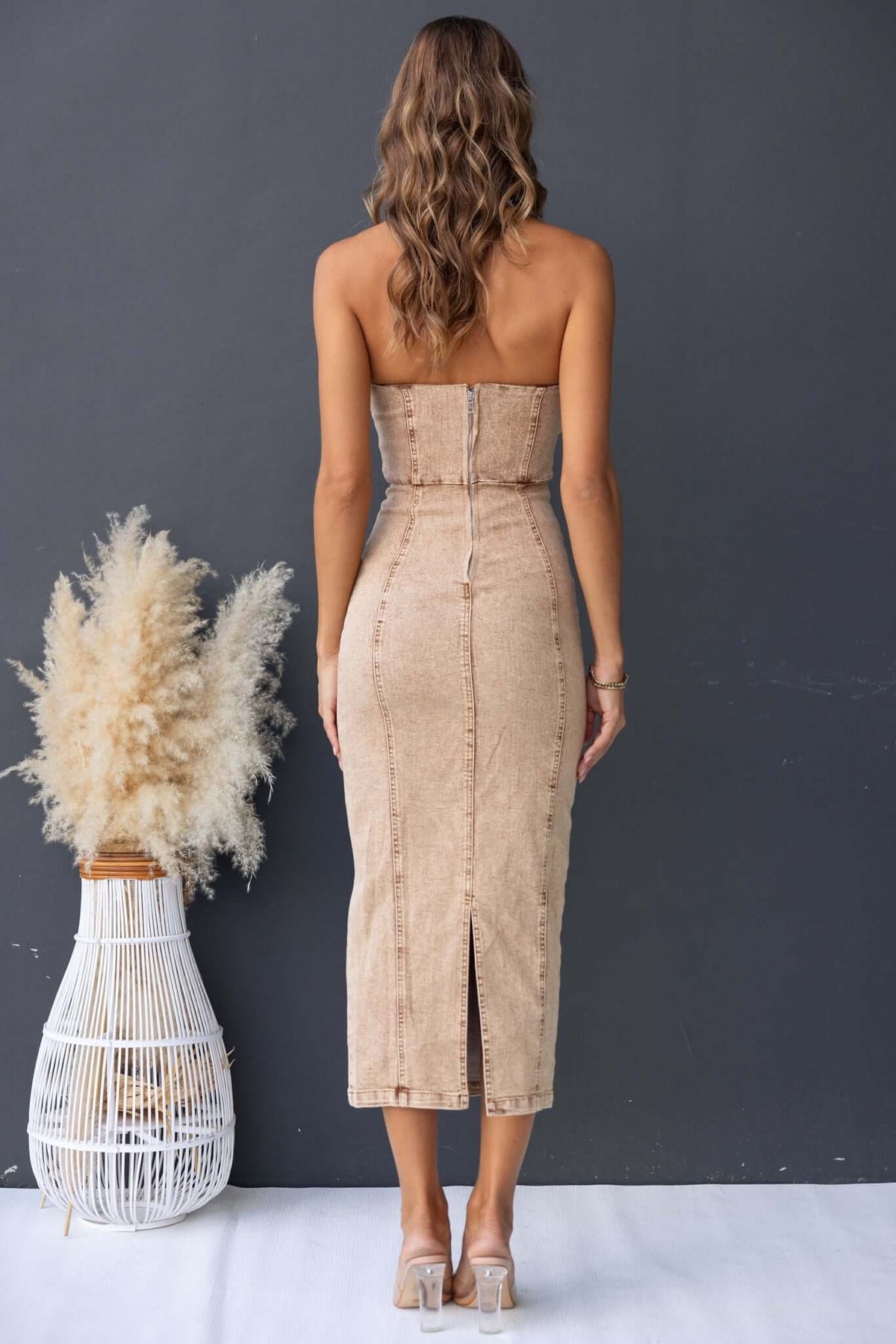 Back view of Varney Denim Midi Dress featuring strapless design and back split, displayed with a decorative plant.