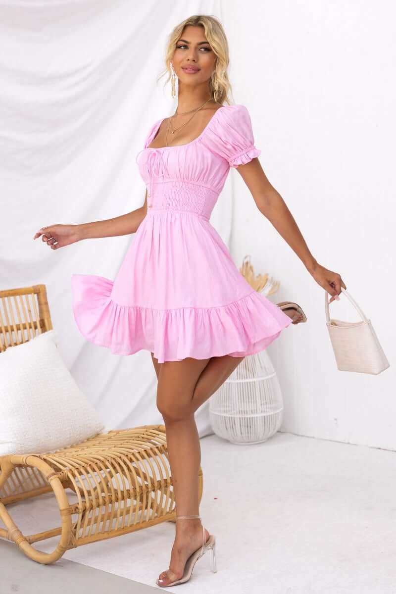 Model wearing the Love In Motion Dress in pink, featuring a square neckline and tiered skirt, styled with clear heels.