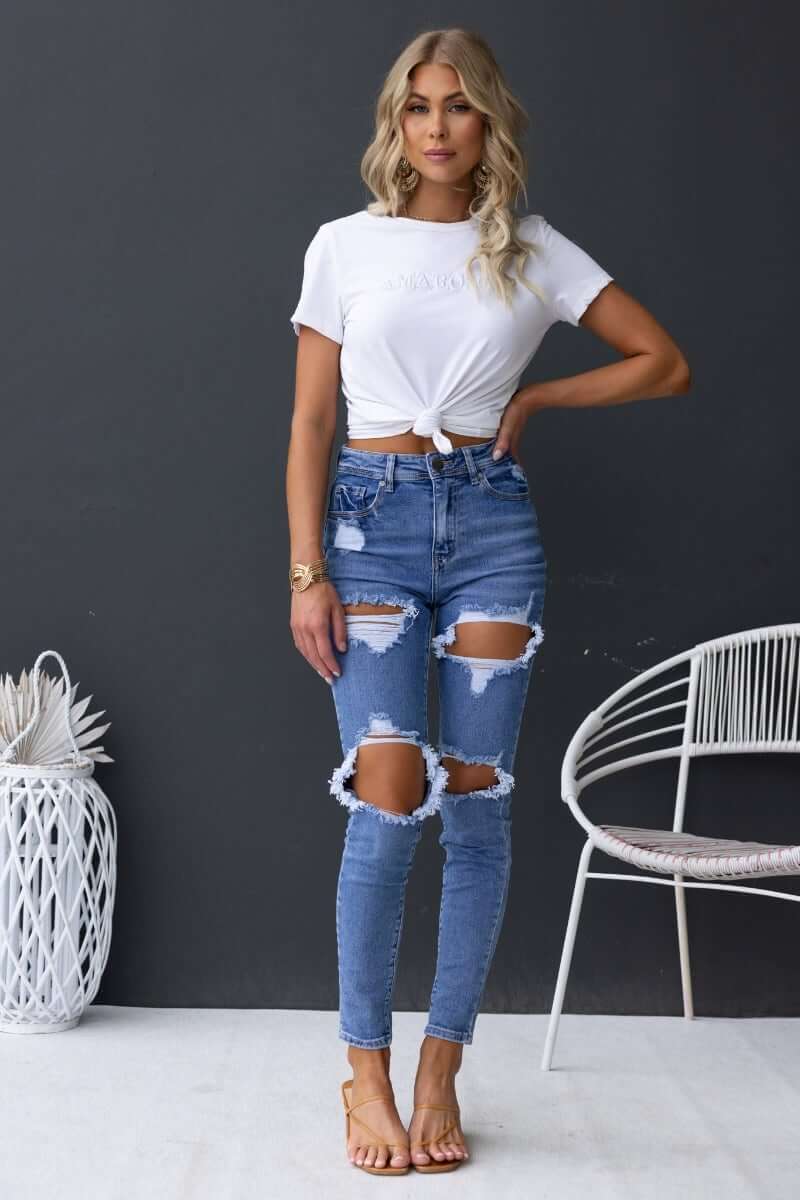 Model wearing distressed Fletcher Jeans with mid-high waist and rips, paired with a white knotted tee, stylish and casual outfit.