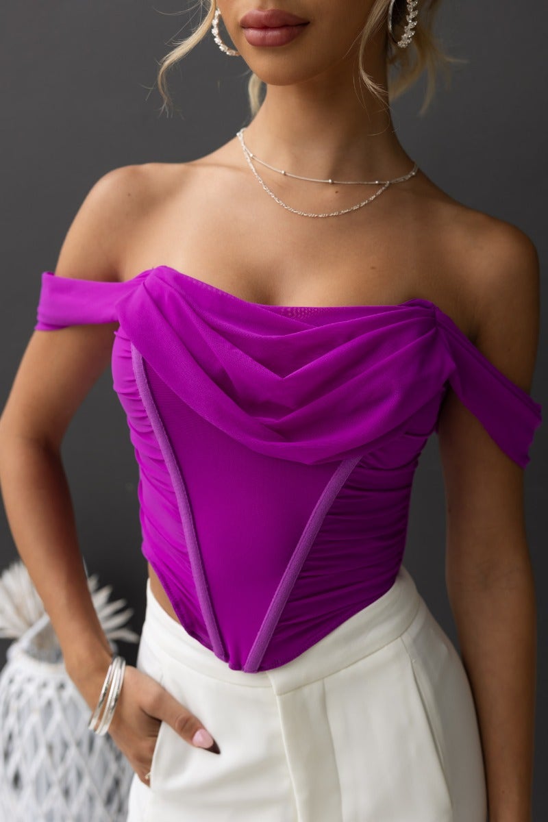 Model wearing Taria Top, a purple corset off-shoulder top with boning, paired with white pants, showcasing its stylish design.