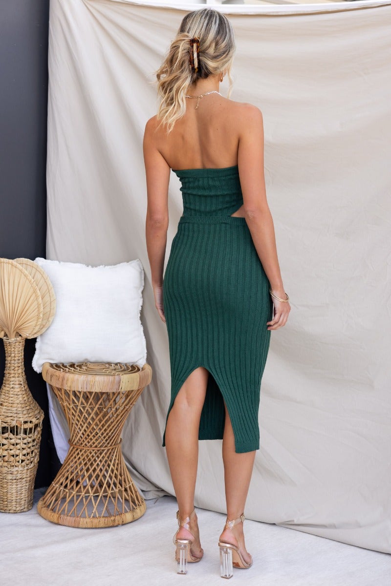 Back view of a model wearing a ribbed knit green midi dress with a cut-out waist and a back slit.
