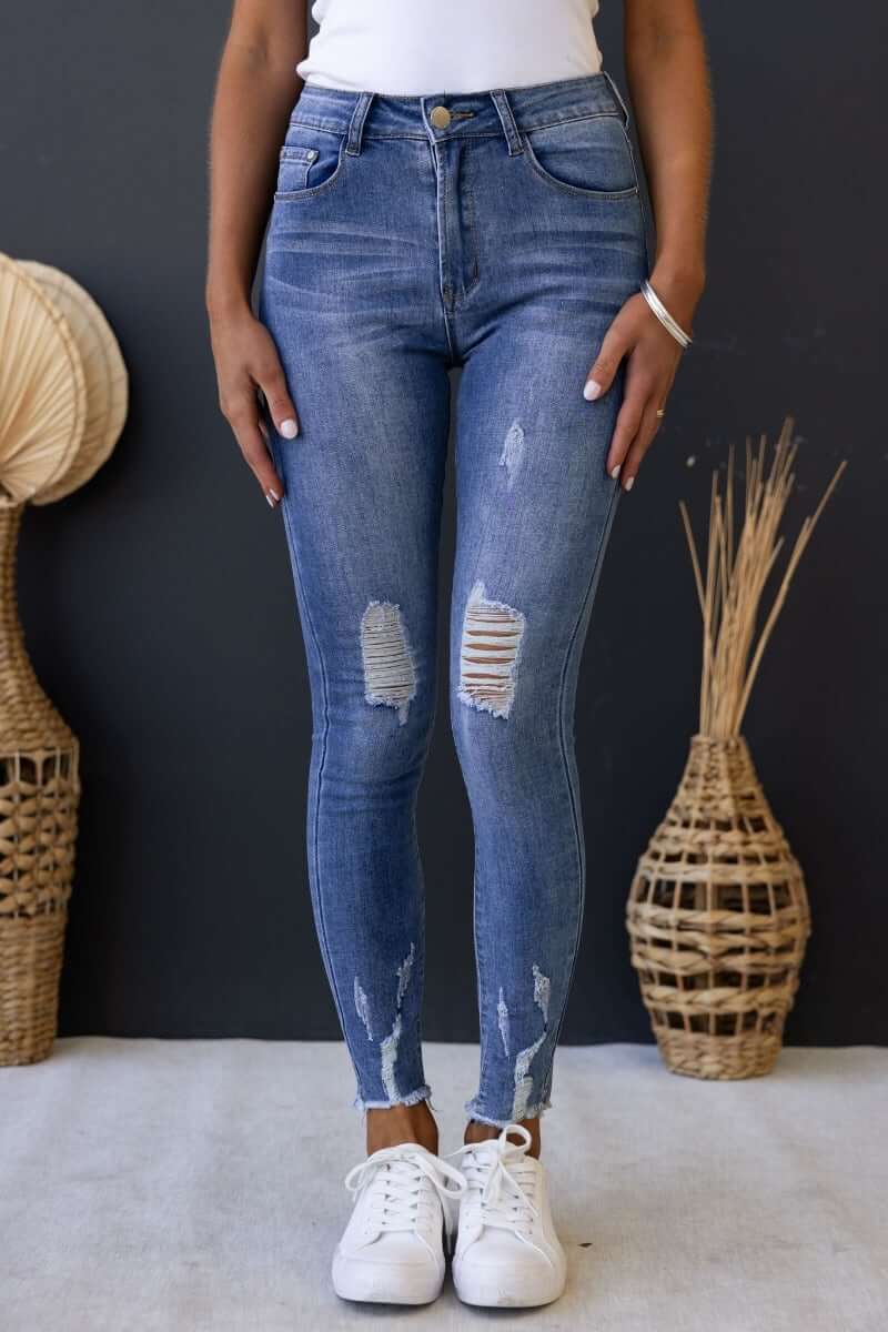 Qilli Jeans in blue denim with distressed rips and raw hem, styled with white sneakers against a textured backdrop.