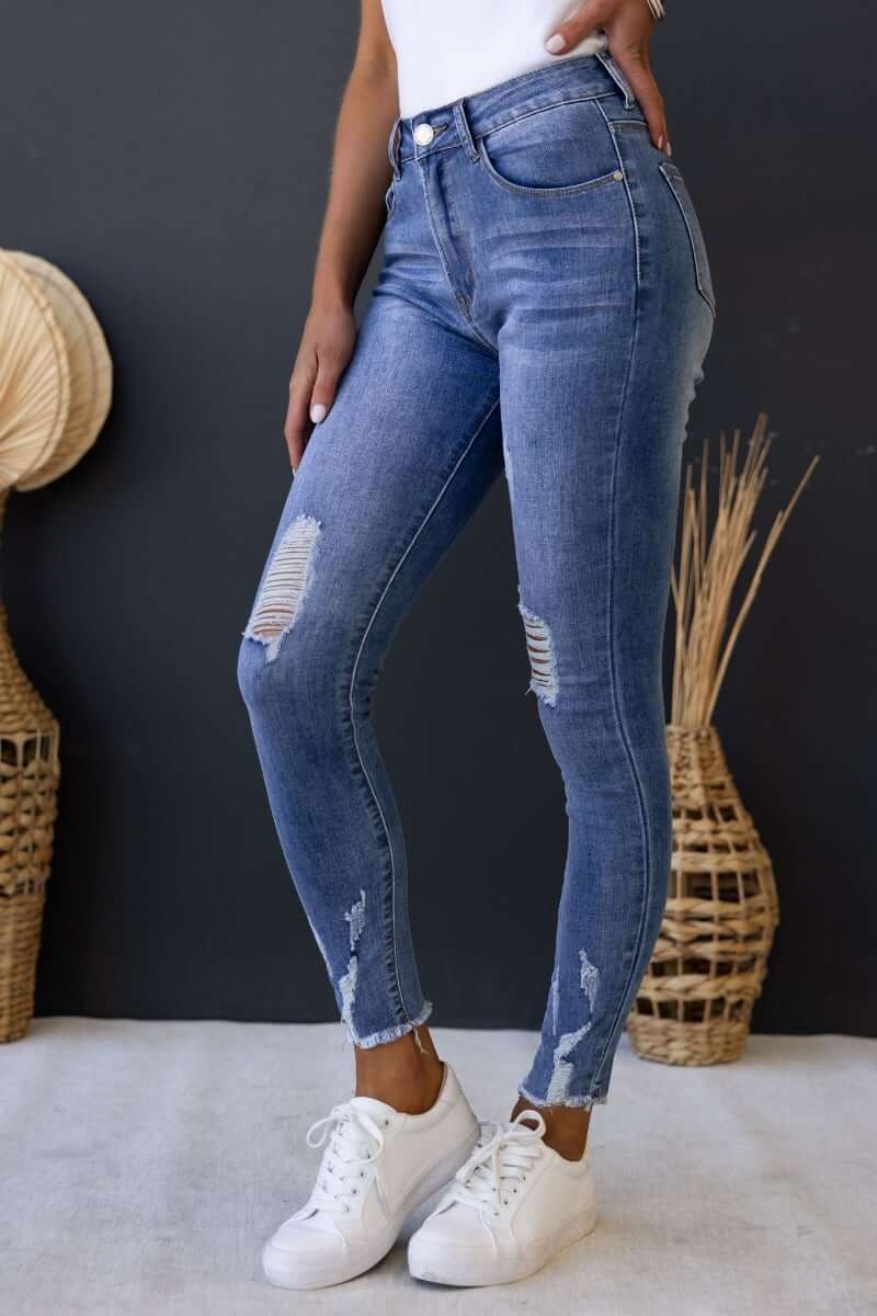 Model wearing Qilli distressed denim jeans with ripped details and raw hem, styled with white sneakers.