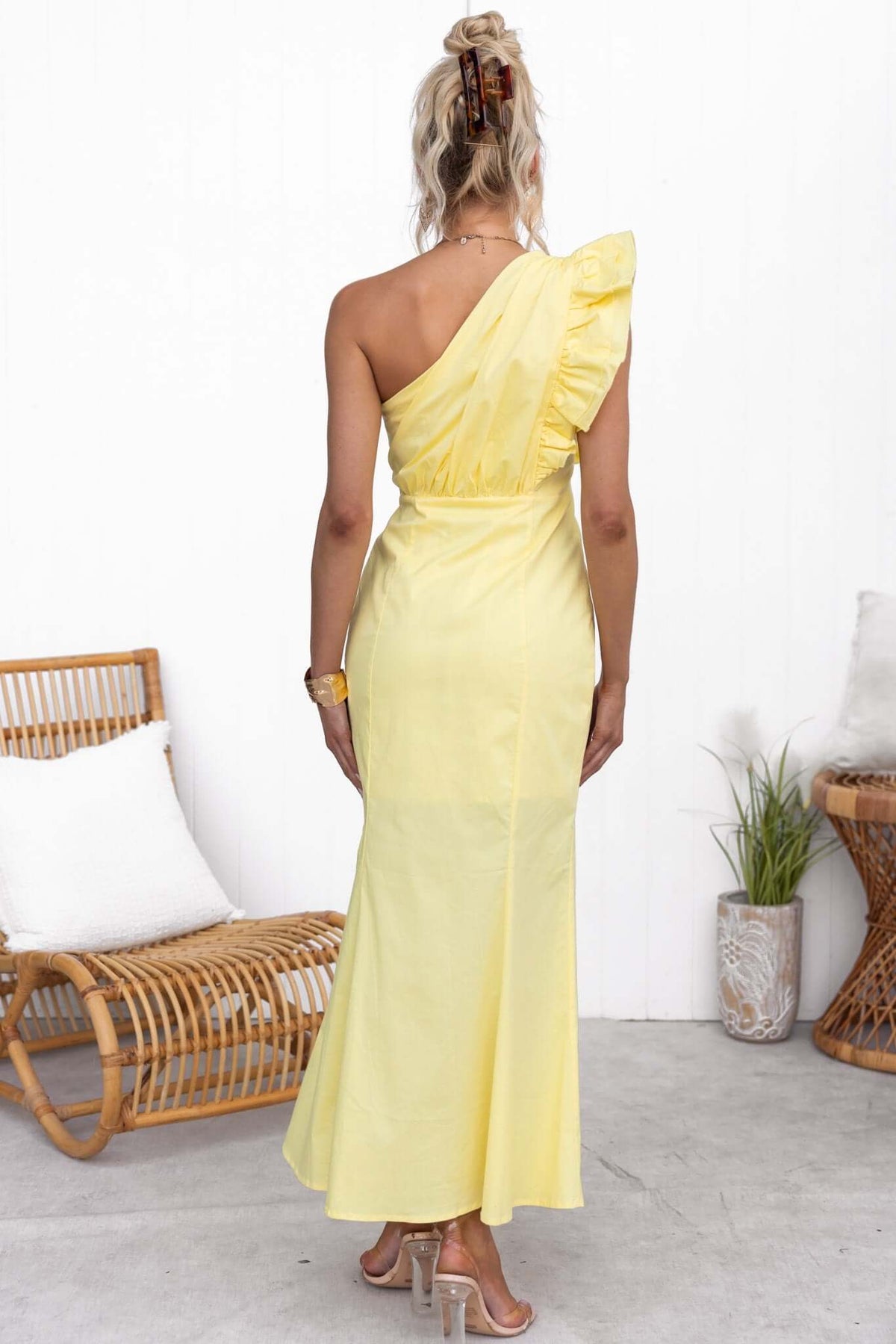 Back view of a woman in a one-shoulder yellow maxi dress with ruffle detail, styled in a modern interior.