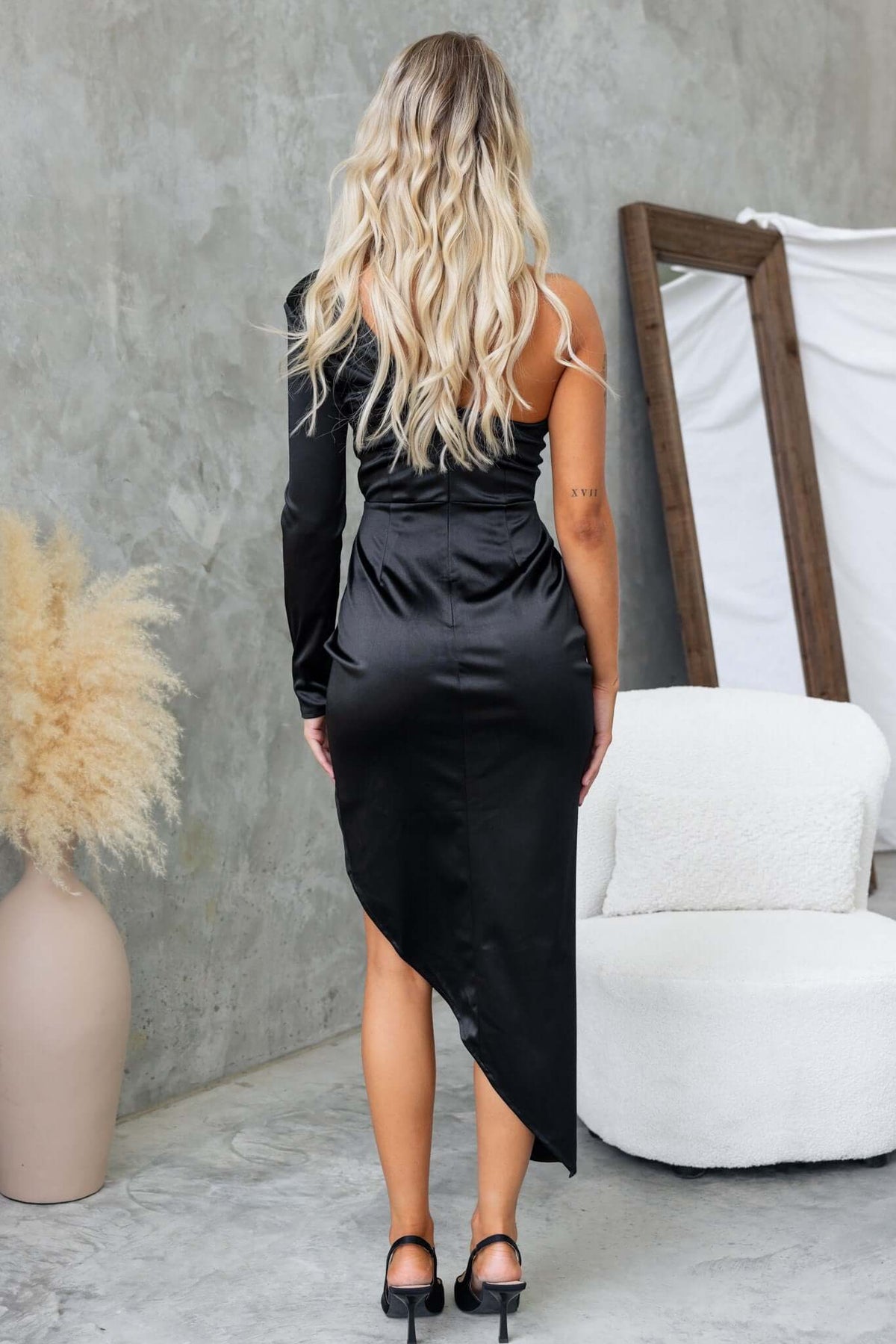 Back view of a woman wearing a black one-shoulder Rommey maxi dress with asymmetric hemline and satin finish.