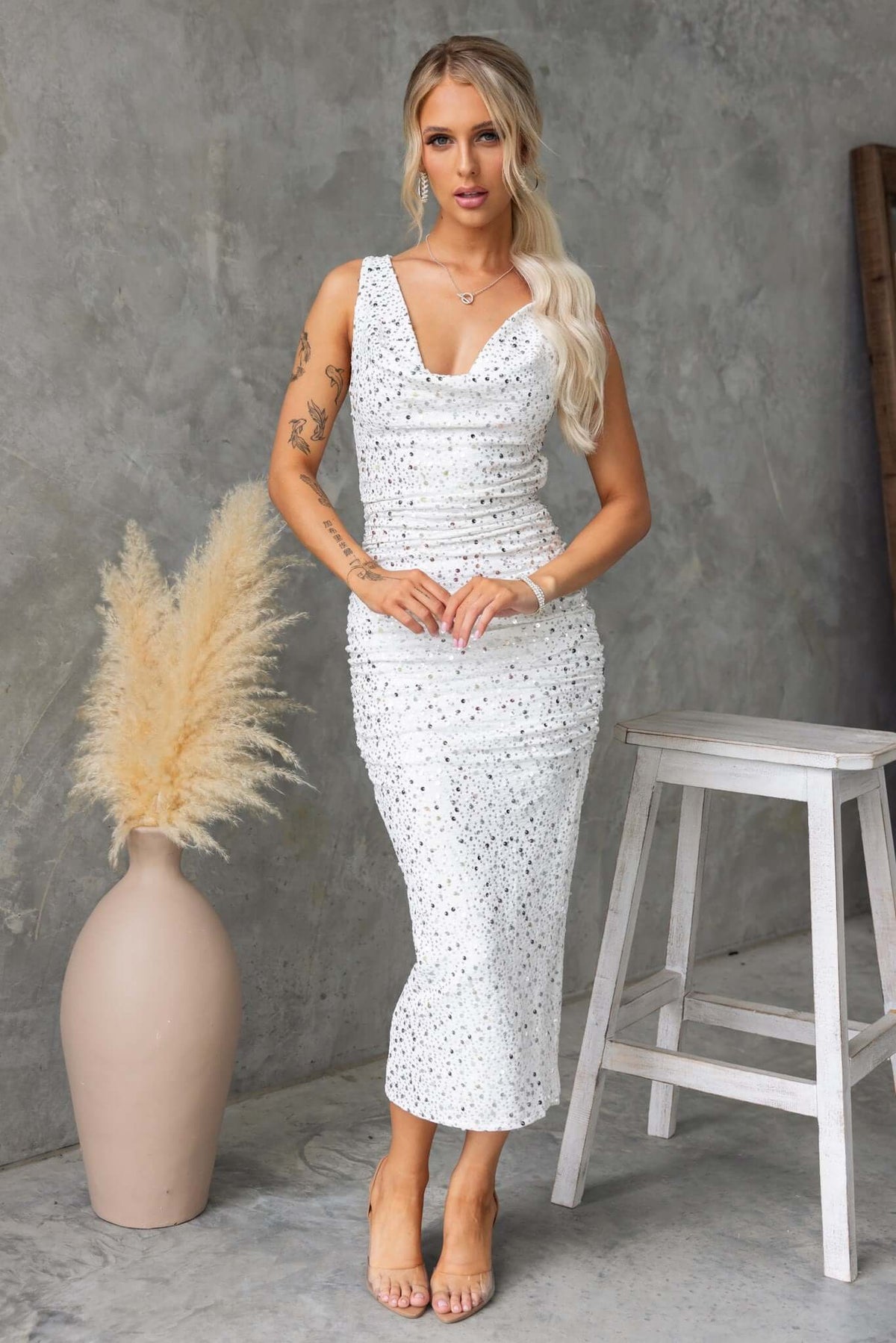 Model wearing a white sequined midi dress with a cowl neckline, styled against a gray backdrop and a cozy ambiance.