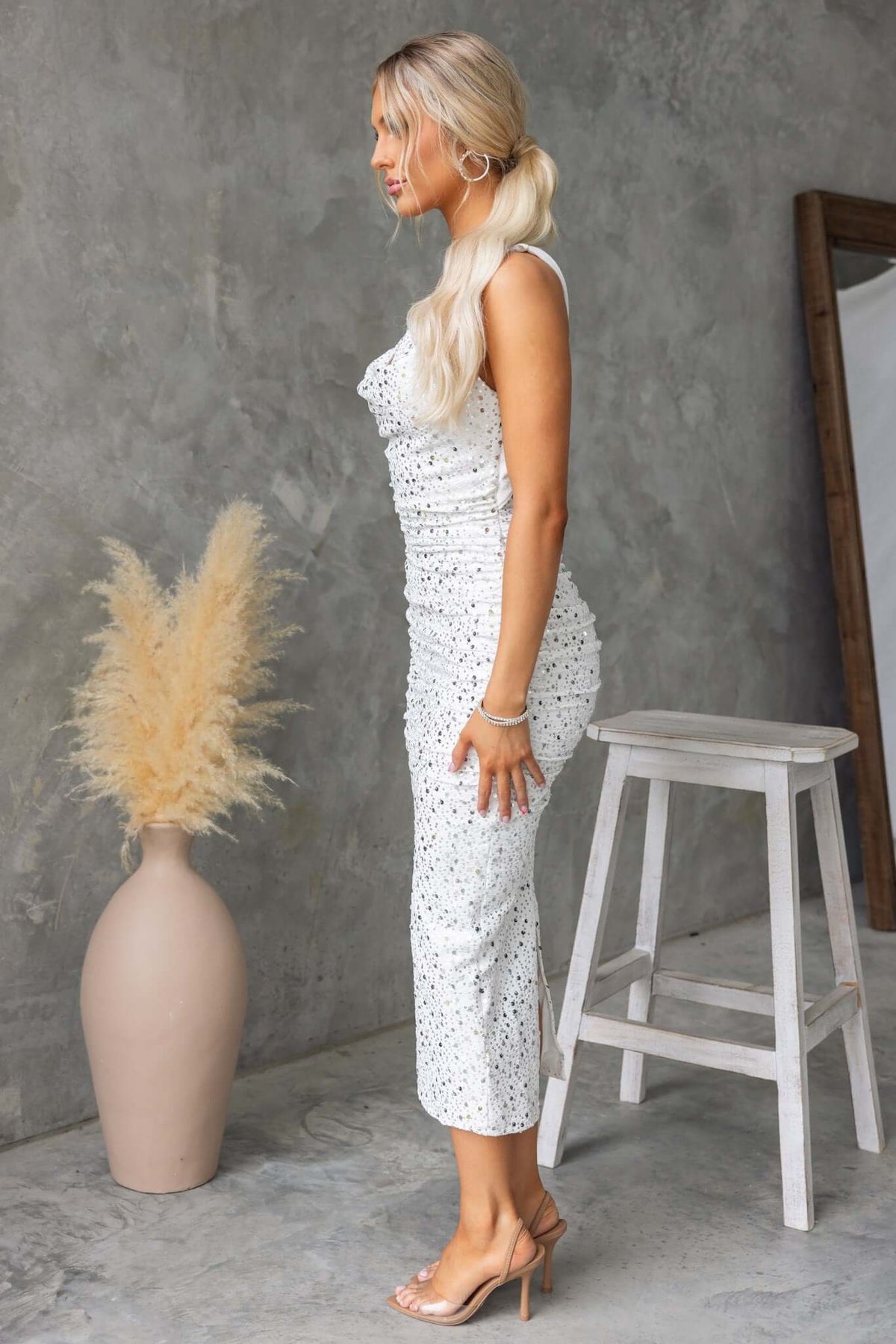 Model wearing an elegant white sequined dress with ruching, styled with a ponytail and standing next to a vase.