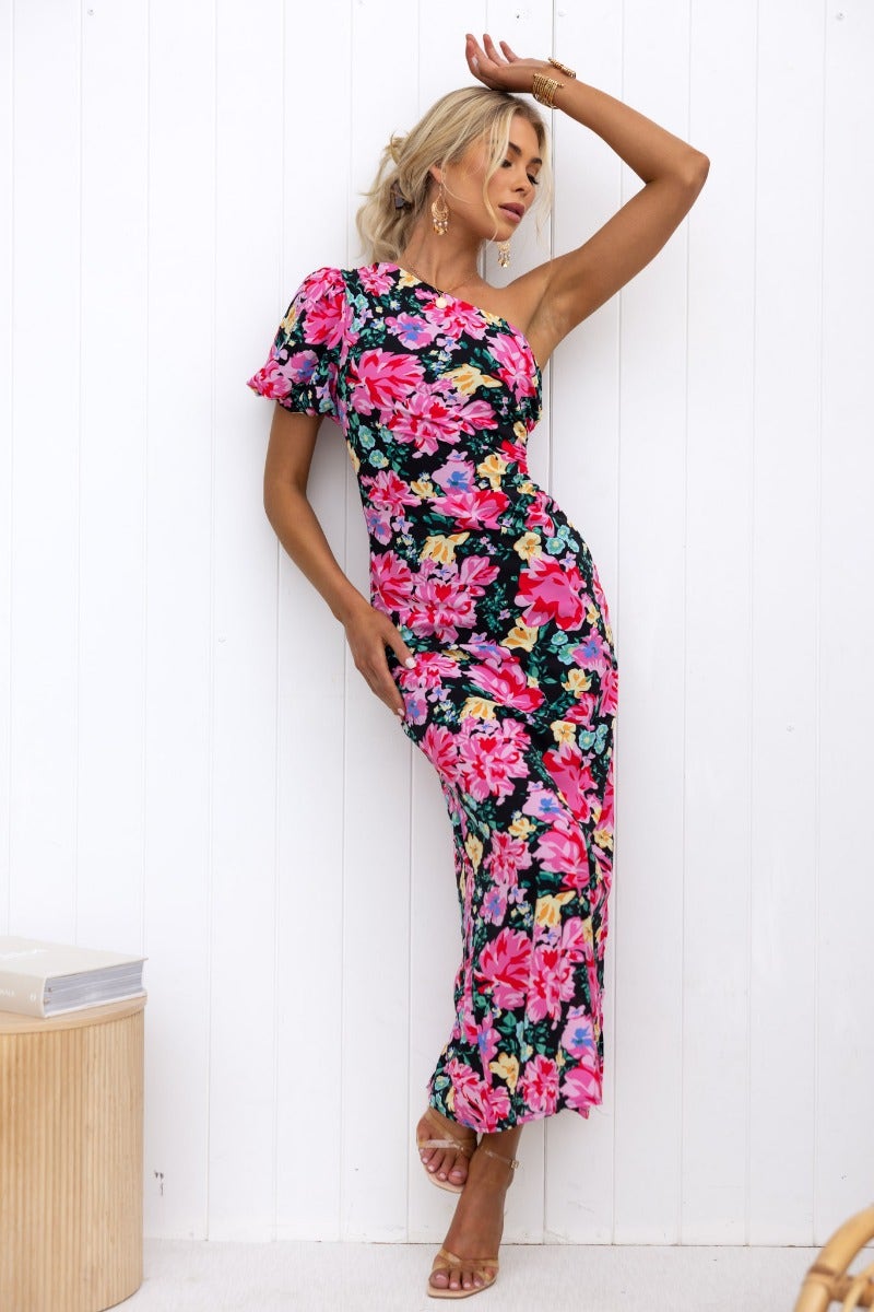 Woman wearing a black floral print one-shoulder maxi dress with side split and ruching, perfect for summer events.