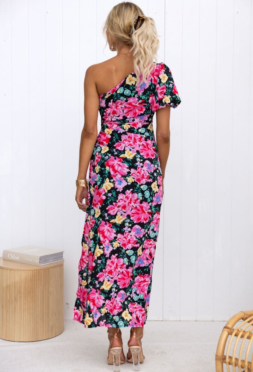 Back view of Karana Maxi Dress showcasing vibrant floral print on black, one-shoulder design with side split.