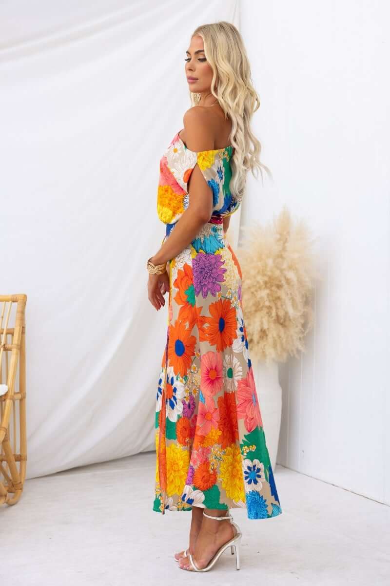 Model in colorful floral maxi dress with off-shoulder design, showcasing vibrant flowers and a side split.
