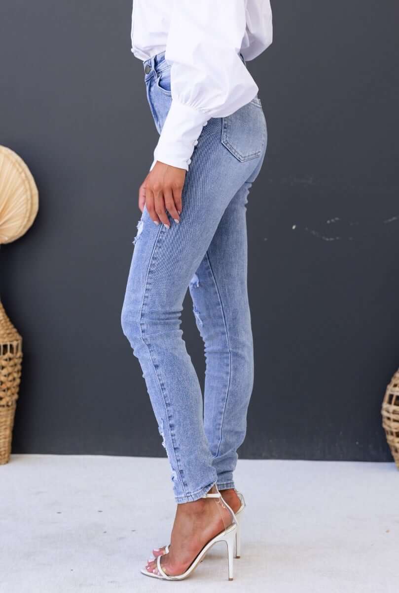 Side view of Mecka Jeans, mid-high waist denim with distressed rips, styled with a white blouse and heels.