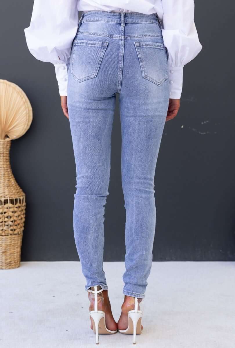 Rear view of Mecka Jeans with mid-high waist, skinny fit, distressed rips, and light blue denim, styled with heels.