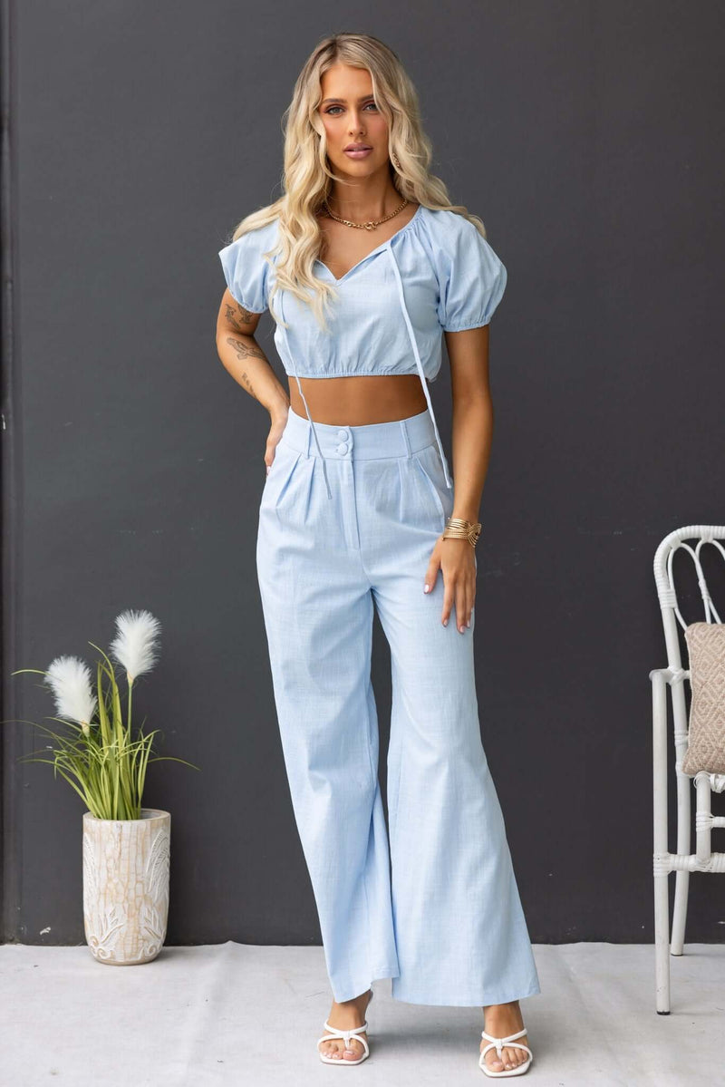 Model wearing Socorro Top in light blue, featuring a tie at bust and elastic sleeves, paired with wide-leg pants.