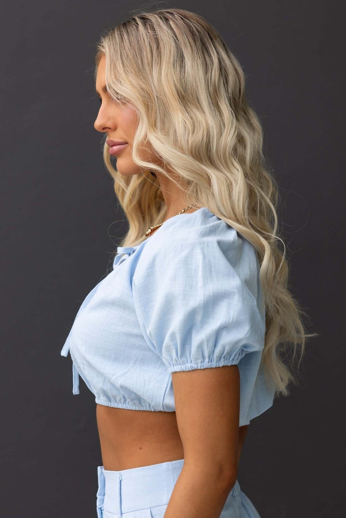 Profile view of a woman in a light blue Socorro Top with tied bust and puff sleeves, showcasing a stylish summer look.