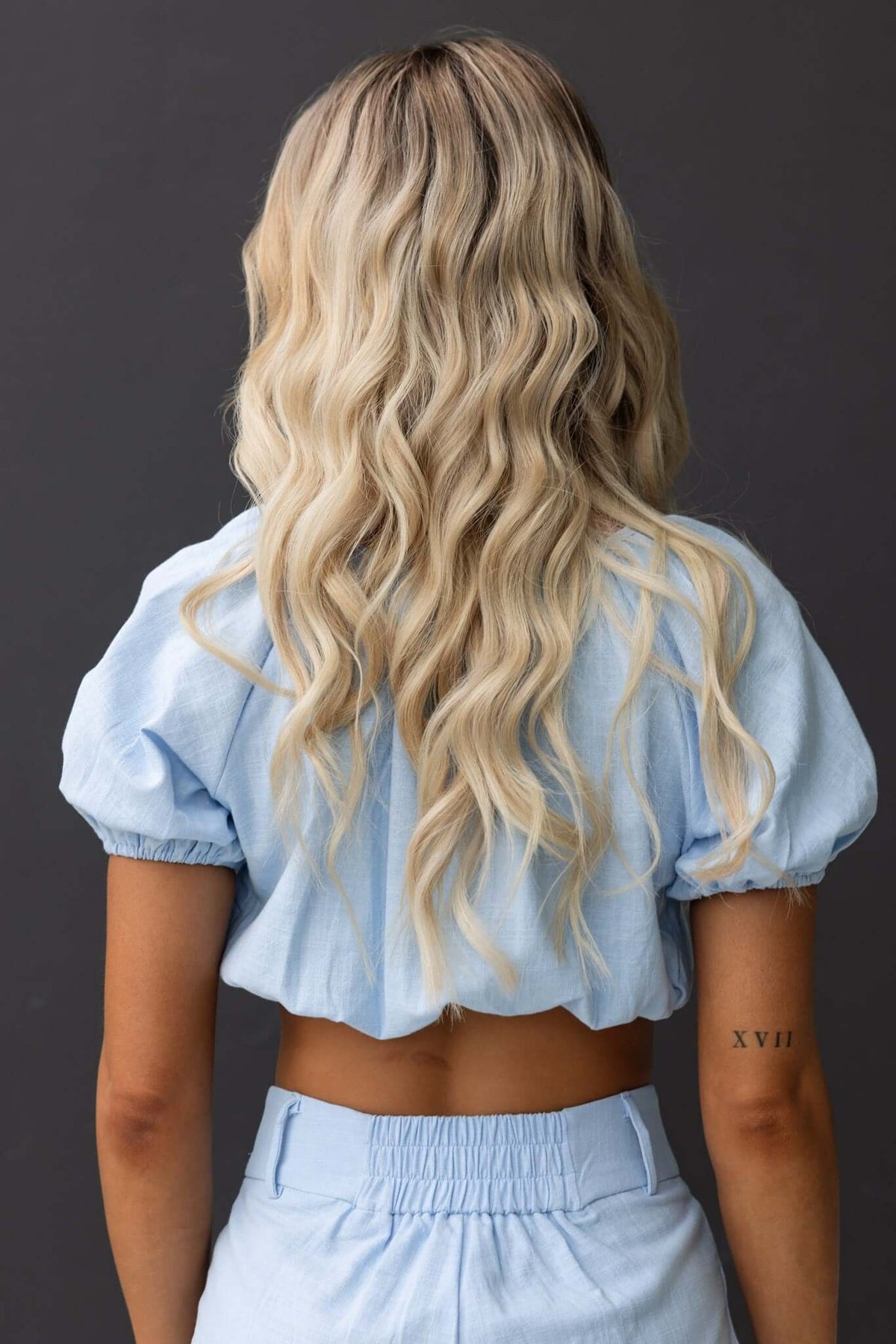 Back view of a woman in a light blue cropped top with puff sleeves and a tie at the bust, showcasing wavy blonde hair.