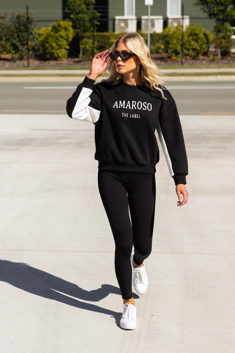 Model walking in black Amaroso jumper with white sleeves, styled with black leggings and white sneakers.