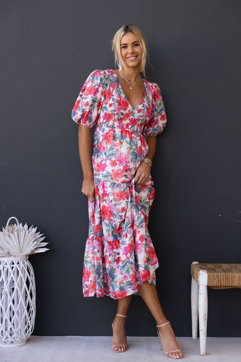 Model wearing Ave Maxi Dress, a floral print dress with V-neckline and puff sleeves against a dark wall.