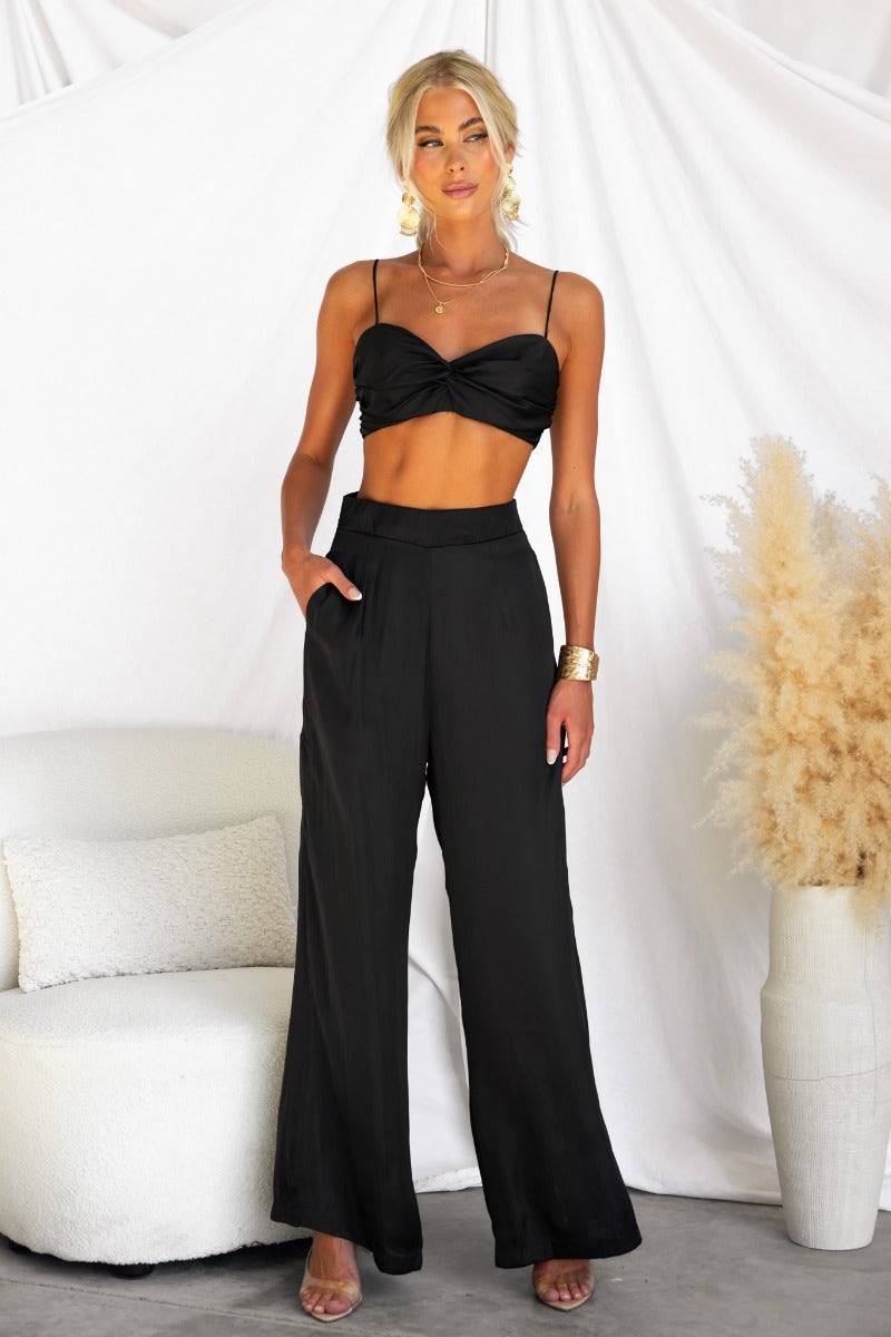 Model wearing a black satin Show Stopper Top with thin straps, paired with black wide-leg pants.