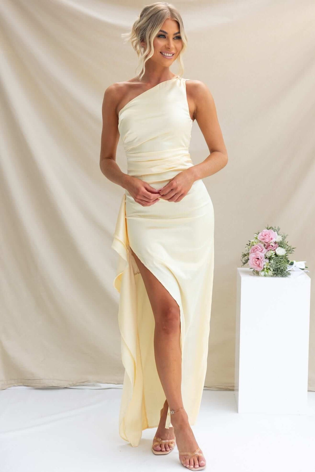 Model wearing Dimmi Maxi Dress, a one-shoulder satin gown with a thigh-high slit and ruffle trim.