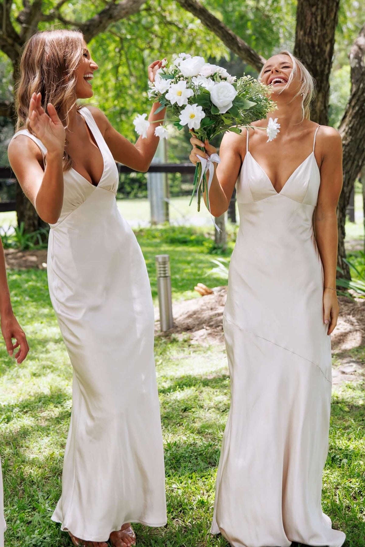Two bridesmaids in elegant Coralina maxi dresses laughing and holding flowers in a sunny outdoor setting.