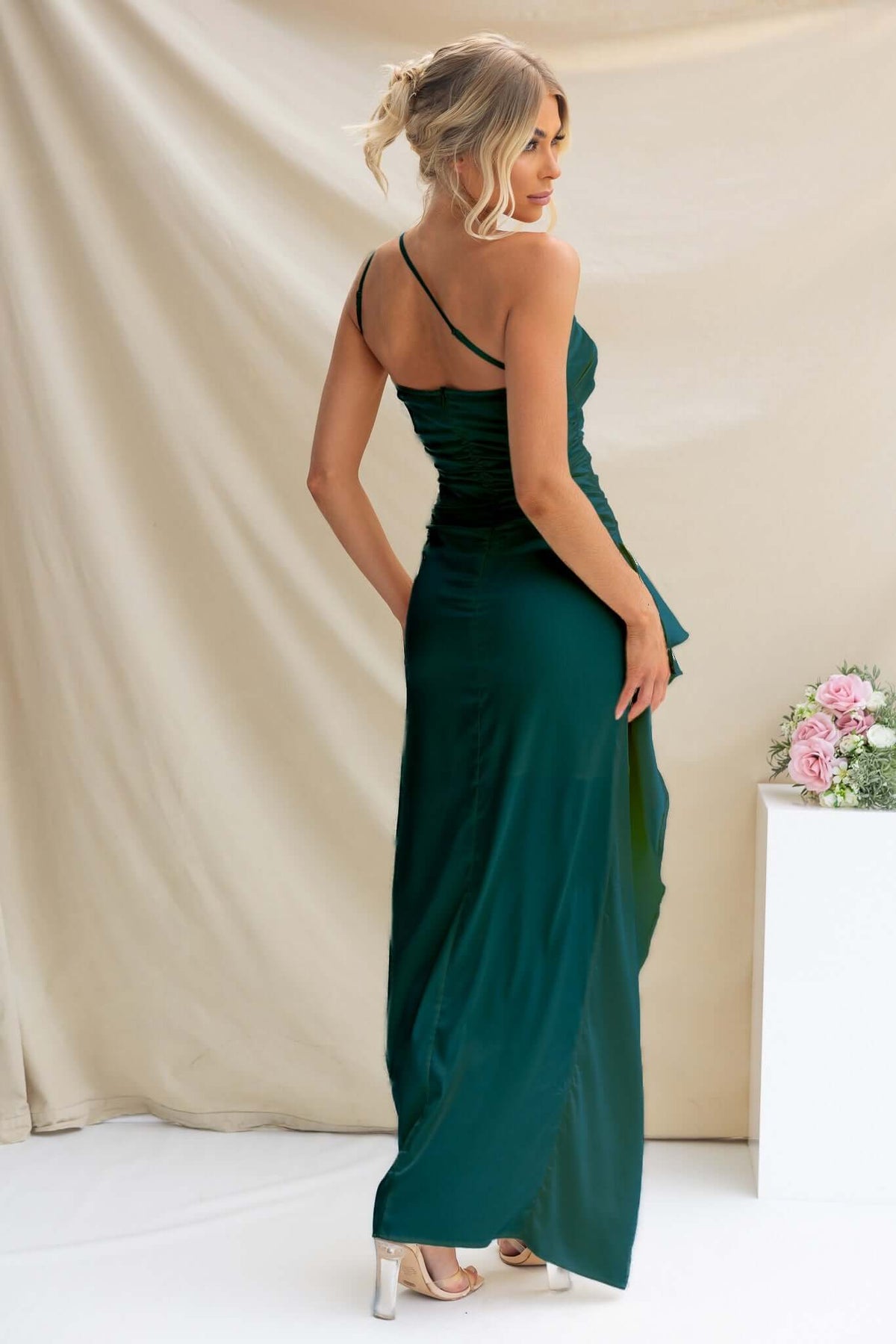 Back view of the Dimmi Maxi Dress in deep green, showcasing satin fabric and an asymmetrical hemline with ruffle trim.