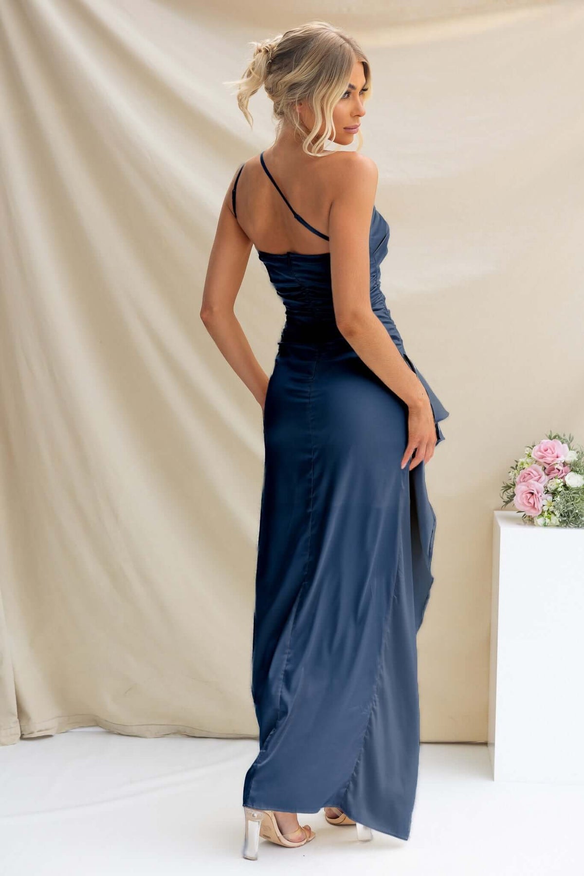 Woman in a navy satin Dimmi Maxi Dress, featuring a one-shoulder neckline and ruffle trim, showcasing the elegant back design.