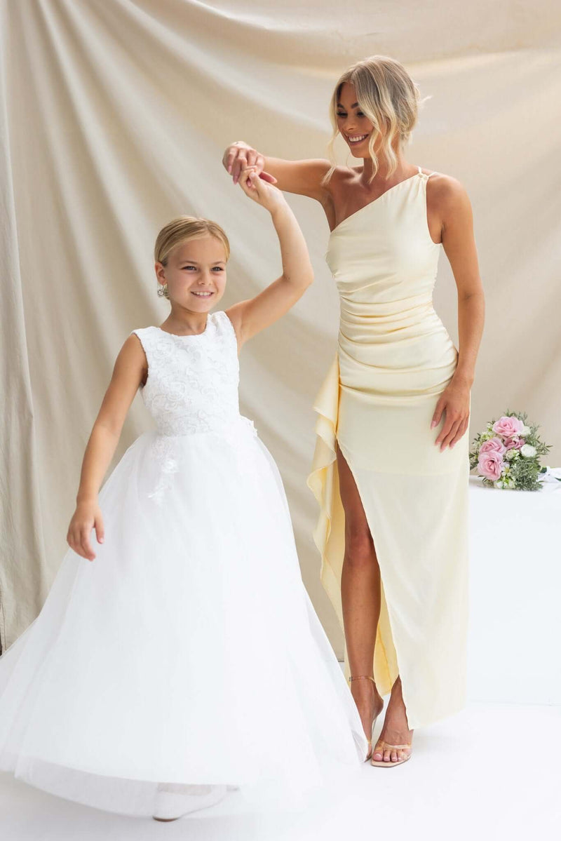 A woman in a satin one-shoulder maxi dress with a girl in a white dress, dancing together in a soft, elegant setting.