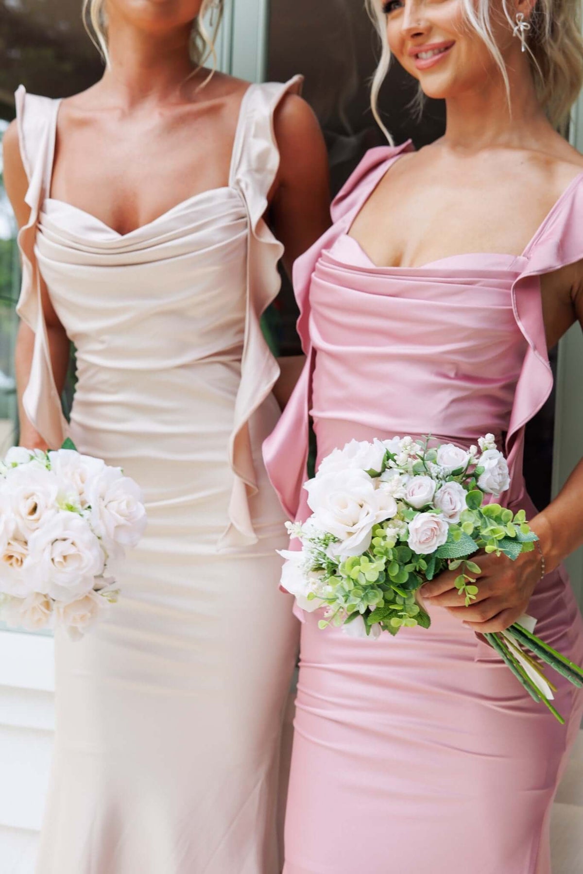 Two women in elegant maxi dresses, one in beige and the other in pink, holding floral bouquets.