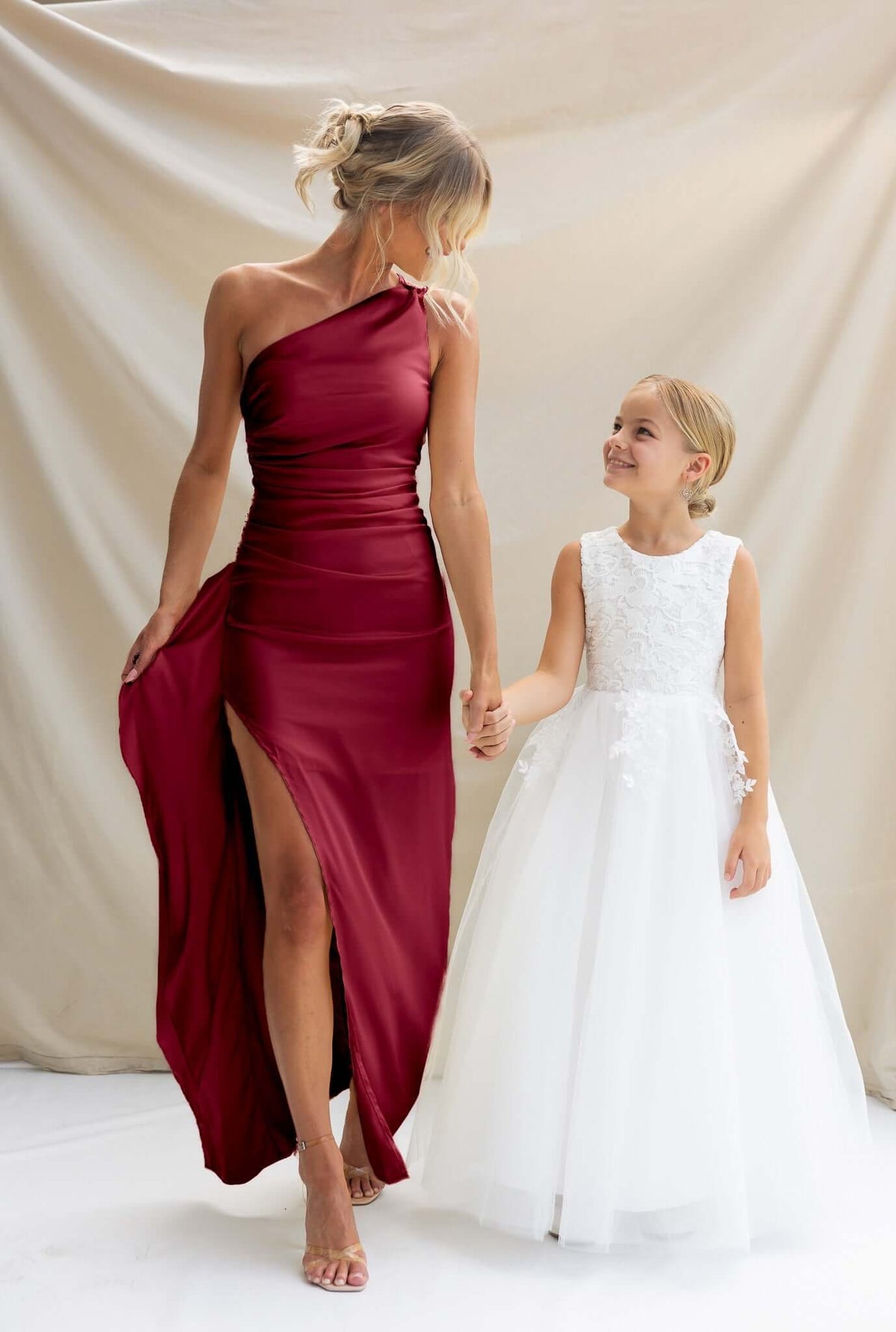 Elegant woman in a one-shoulder burgundy satin maxi dress holding hands with a girl in a white floral dress.