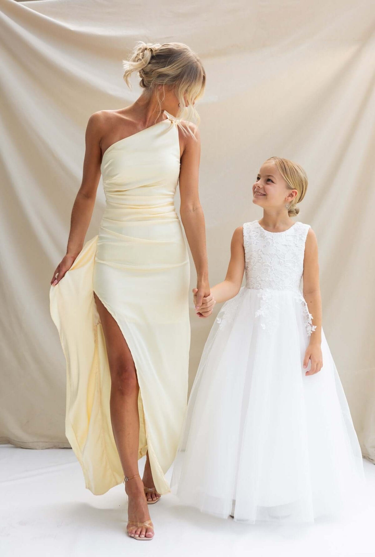Woman in a satin one-shoulder maxi dress holding hands with a young girl in a white floral dress.