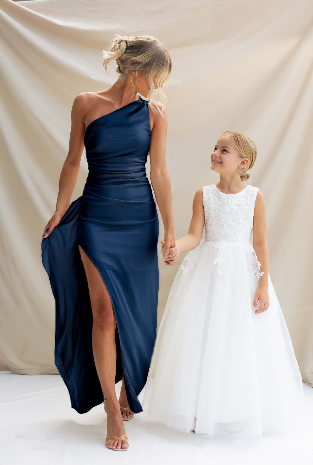 Stylish model in navy satin Dimmi Maxi Dress with one-shoulder neckline, holding hands with a girl in a white dress.