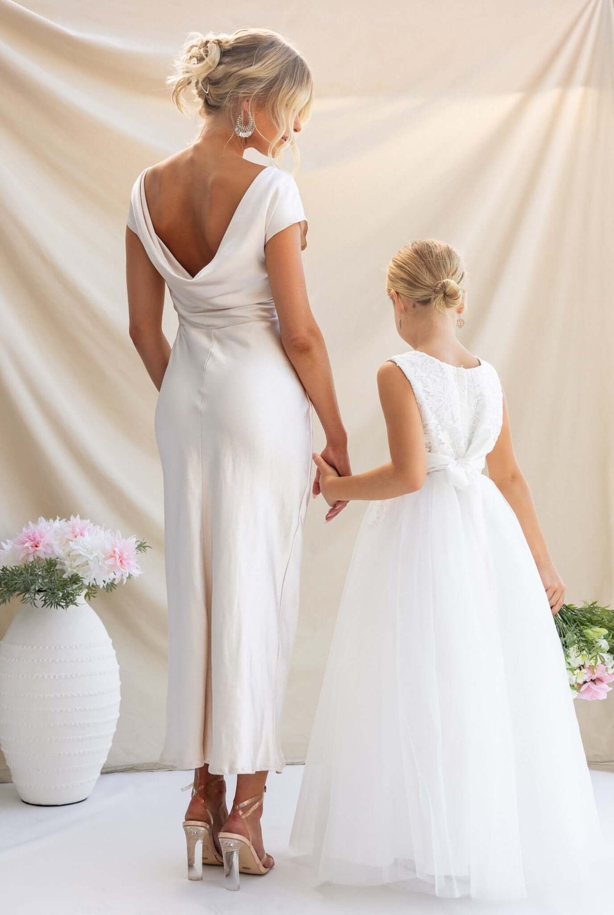 Woman in satin Esmeralda maxi dress with boat neckline holding hands with a girl in a white dress, showcasing elegant bridal style.