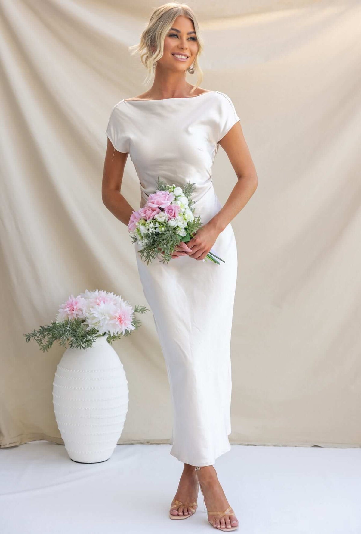 Elegant woman in a satin Esmeralda Maxi Dress holding a bouquet, showcasing a boat neckline and draped back.