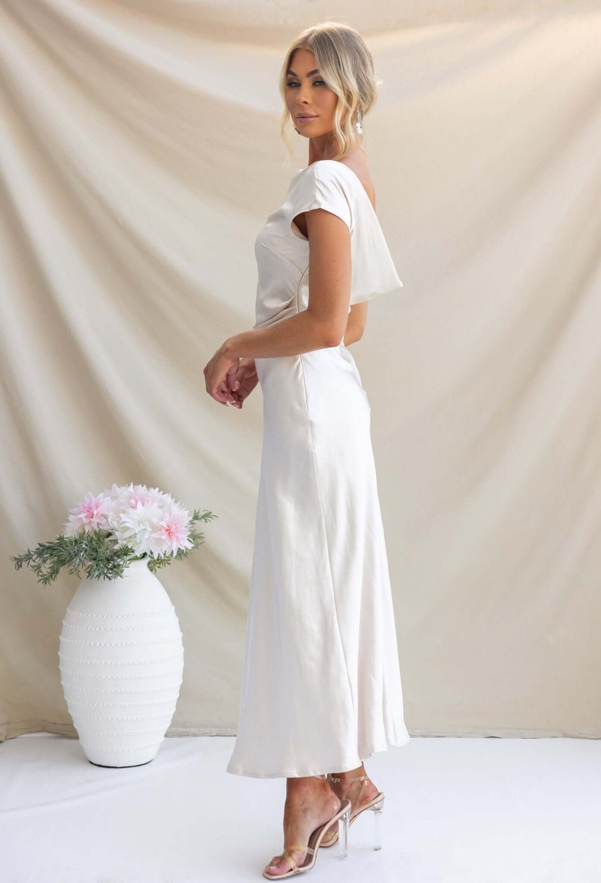 Model showcasing the Esmeralda Maxi Dress with boat neckline and draping details, styled with a potted plant backdrop.