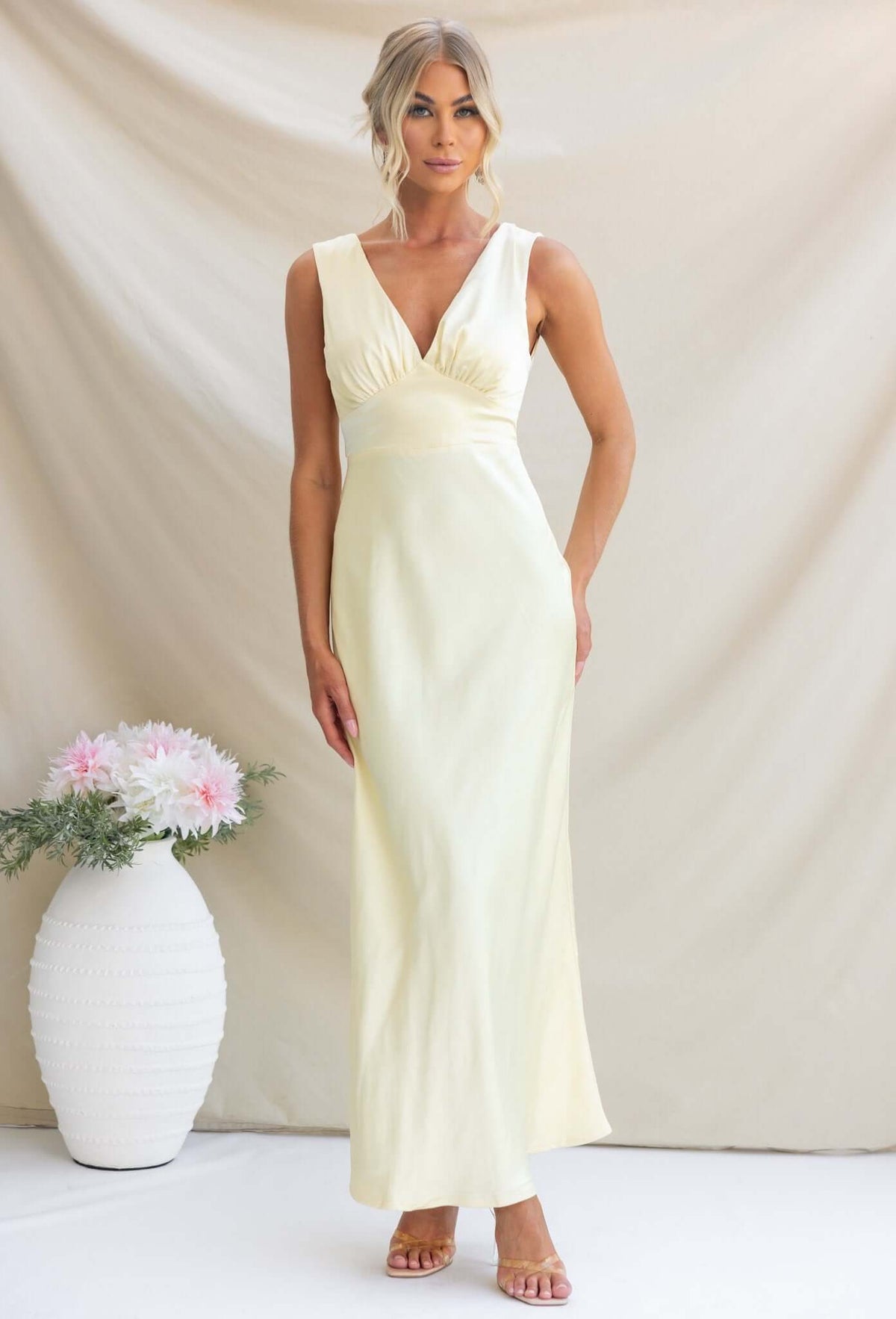 Alika Maxi Dress in soft yellow satin with V-neckline and back split, modeled by a woman in a minimalistic setting.
