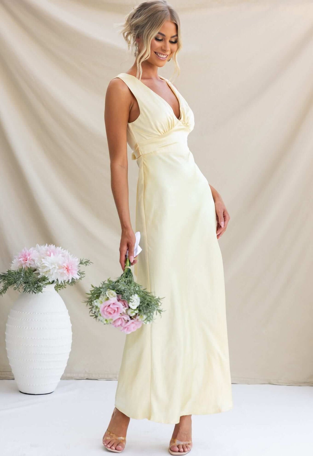 Model wearing Alika Maxi Dress in yellow with V-neckline, tied waist, and floral bouquet.