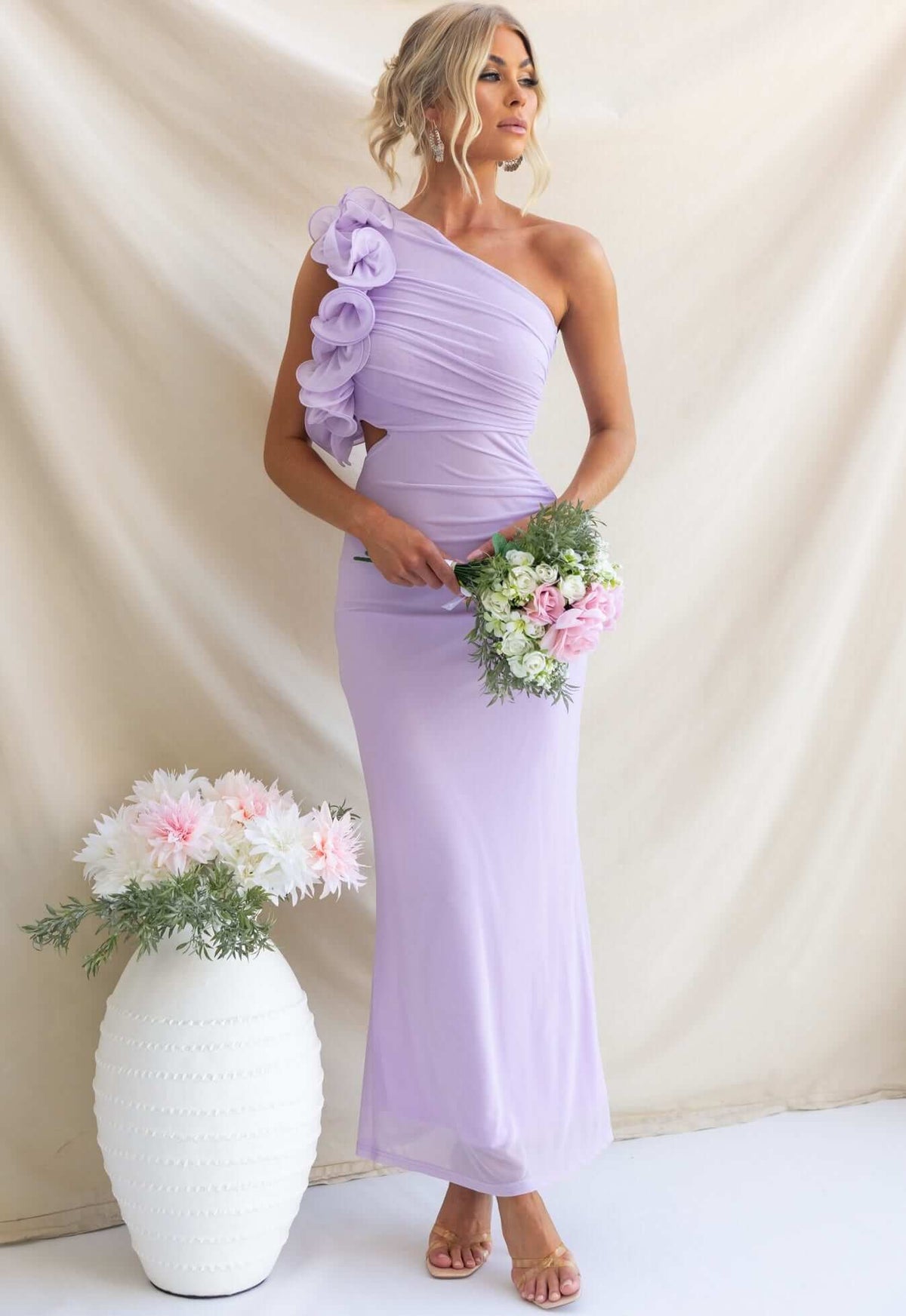Model wearing a lavender Meriden Maxi Dress with one-shoulder design and floral detailing, holding a bouquet of flowers.