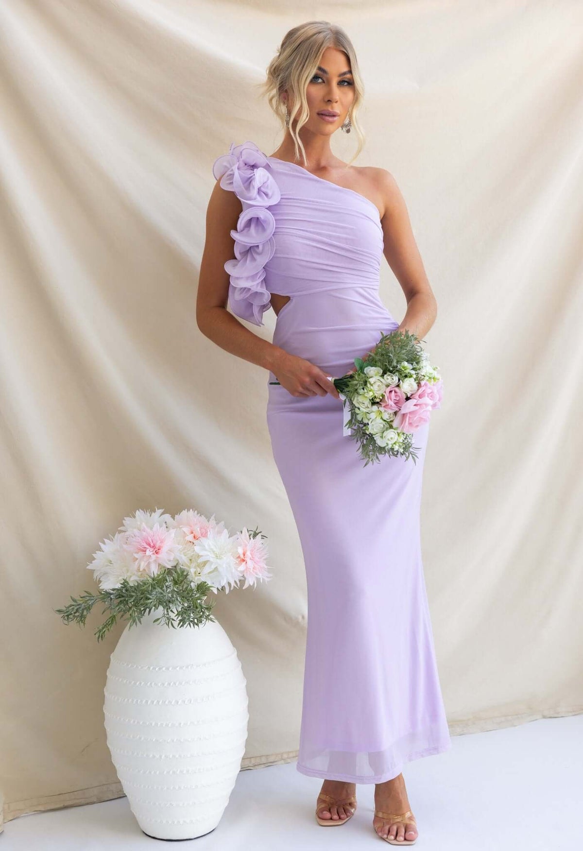 Model in a lilac Meriden Maxi Dress featuring a one-shoulder neckline and floral details, holding a bouquet of flowers.
