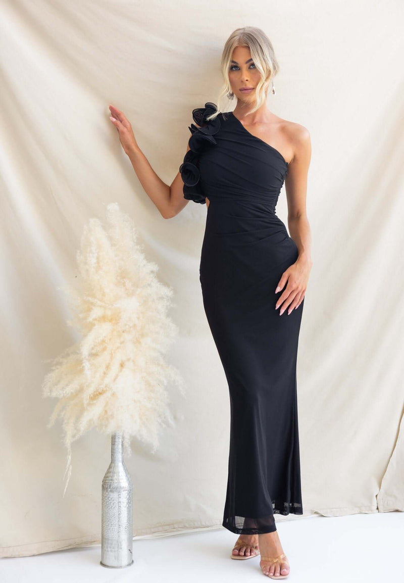 Model in a black Meriden Maxi Dress featuring a one-shoulder neckline and floral detail against a neutral background.