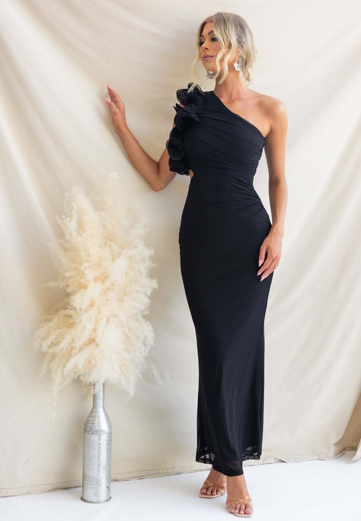 Model wearing a black Meriden Maxi Dress featuring a one-shoulder design and floral detail, styled with a vase and pampas grass.